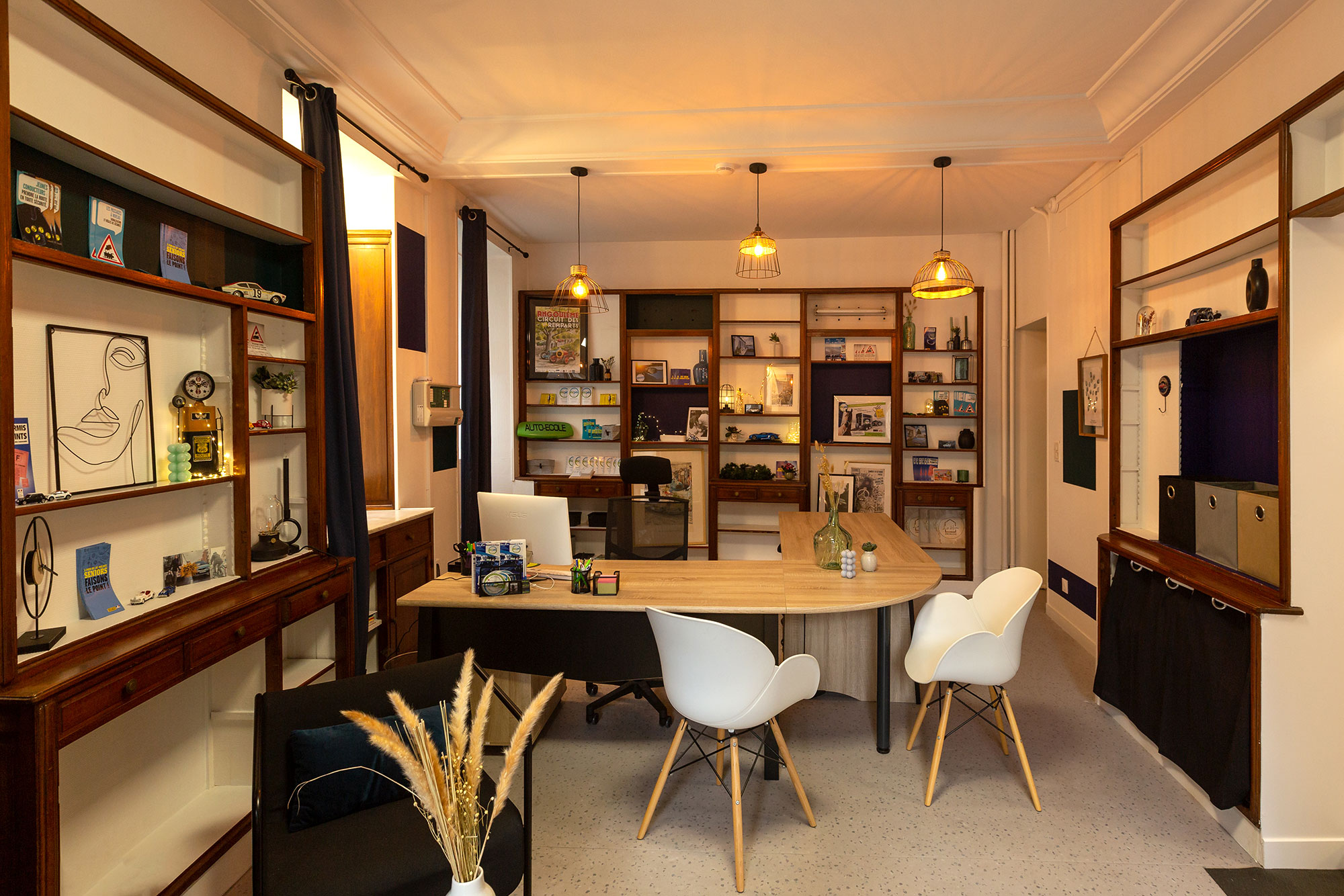 Bureau aménagé avec rangements muraux en bois, table de travail centrale, chaises design blanches, étagères décorées, éclairage suspendu et espace détente avec fauteuil.