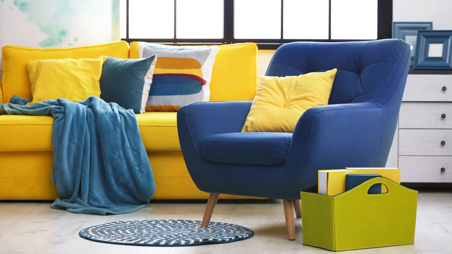 Salon coloré avec canapé jaune vif, plaid bleu, fauteuil bleu marine, coussins jaunes et décorations assorties, tapis rond à motifs et rangement jaune.