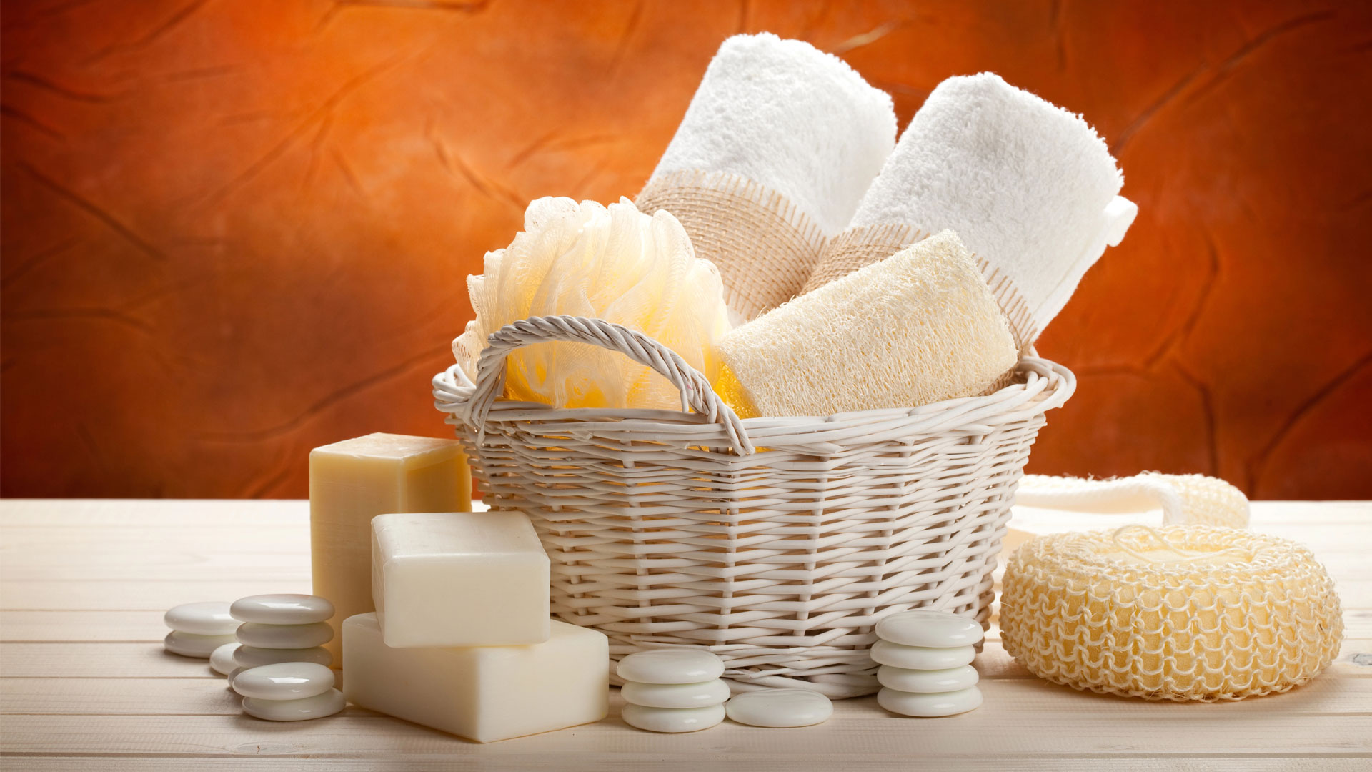 Panier en osier blanc rempli de serviettes, gants de toilette et accessoires de bain, entouré de savons, galets blancs et éponges naturelles sur fond bois clair.